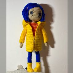 a crocheted doll with blue hair wearing a yellow jacket and red shirt, standing on a white surface