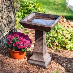 a bird bath sitting next to a tree with flowers in the pot on the ground