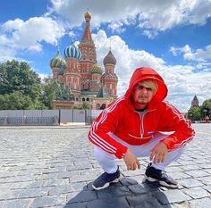 a man in a red hoodie squatting on a brick walkway with a cathedral in the background