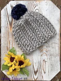 a crocheted hat with a blue pom - pom sits next to a sunflower