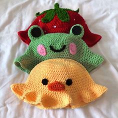 three crocheted hats are sitting on a white sheet, one has a strawberry and the other has a green frog