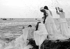 four women in white dresses are standing on rocks near the ocean and splashing water