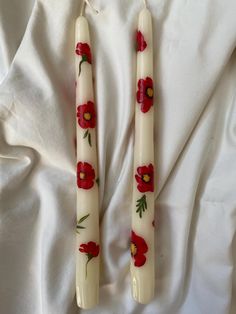 two candles with red flowers on them sitting on a white sheet covered bed, next to each other