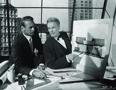 two men in suits look at pictures on a table while another man holds a paintbrush