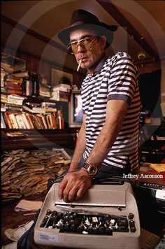 an older man with glasses and a hat standing next to a typewriter