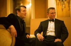 two men in tuxedos are sitting down and one is looking at the camera