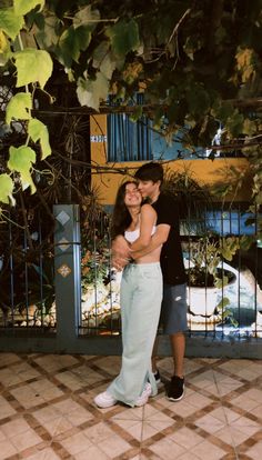 a man and woman standing next to each other in front of a fence with trees