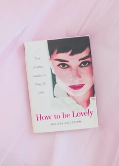 the book how to be lovely is laying on a pink bed sheet with an image of a woman's face