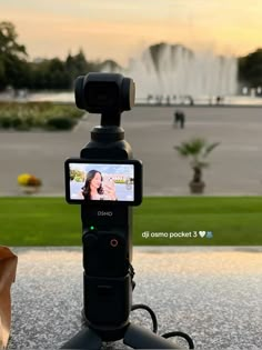 a cell phone is attached to a tripod in front of a fountain with a woman on the screen