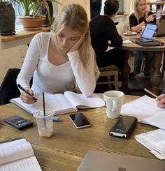 two women sitting at a table with notebooks and papers in front of them, working on laptop computers