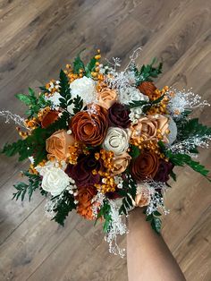 a bridal bouquet with orange, white and brown flowers on a wooden floor in front of a person's hand