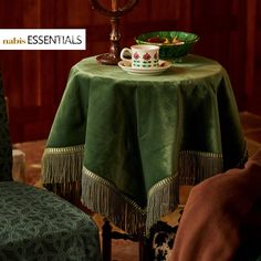 a green table cloth with tassels and a tea cup on it in front of a wooden paneled wall