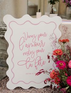 a white sign with pink writing on it next to flowers and a vase filled with water