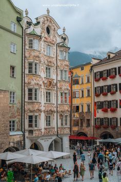 many people are walking around in front of some buildings and tables with umbrellas on them