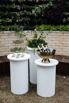 two white pedestals with plants and wine bottles on them in front of a brick wall