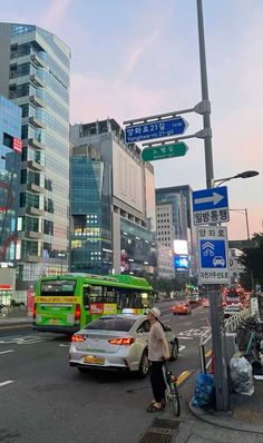 a man riding a bike next to a green bus on a street with tall buildings in the background