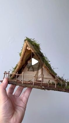 a hand holding up a miniature house made out of wood and mossy material with a roof