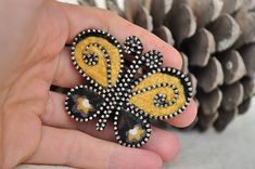 a hand holding a yellow and black flower brooch on top of a pine cone