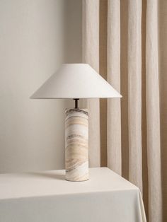 a lamp sitting on top of a white table next to a window with curtains behind it