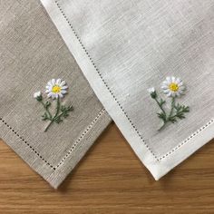 two white linen napkins with embroidered daisies on the side, sitting on a wooden surface