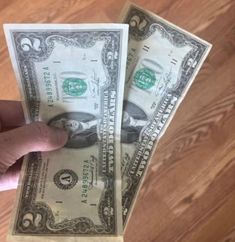 a person holding two stacks of one hundred dollar bills in their left hand, on a wooden floor