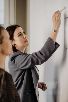 a woman is writing on a whiteboard while another woman looks at the wall behind her