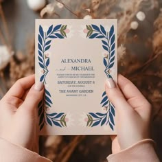 a person holding up a wedding card in front of some dried plants and flowers on the table