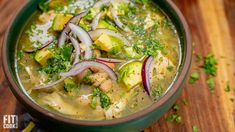 a green bowl filled with soup and vegetables on top of a wooden table next to a spoon