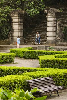 two people are walking in front of a mazed area with a bench and trees