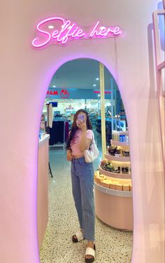 a woman taking a selfie in front of a pink neon sign that says selfie here