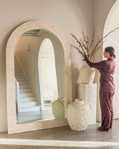 a woman standing in front of a large mirror next to a vase with flowers on it