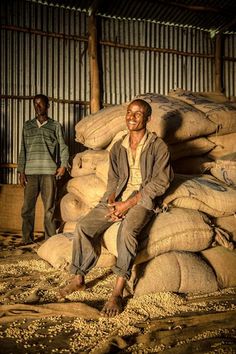 two men standing next to bags of grain in a warehouse area with one sitting on the ground