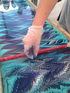 a person with gloves on painting an art piece in blue and green colors, while another person is holding a plastic glove over the top of it