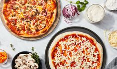 two pizzas sitting on top of a white counter next to other foods and vegetables