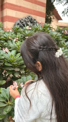 a woman with long hair standing in front of some bushes and flowers looking at her cell phone