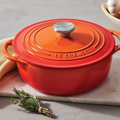 an orange le creuse casserole sits on a cutting board next to garlic