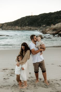 a family standing on the beach in front of the ocean with one holding their baby