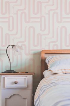 a nightstand with a lamp on top of it next to a bed and wall paper