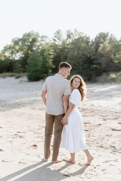 an engaged couple standing on the beach in front of some trees and sand with their arms around each other