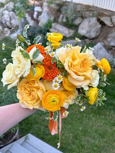 a bridal bouquet with yellow and orange flowers in someone's hand on the grass