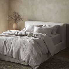 a bed with white linens and pillows in a neutral colored room, next to a vase with dried flowers