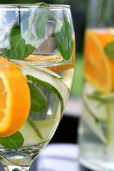 two glasses filled with water, orange slices and green leaves on the rim are sitting next to each other