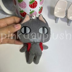 a hand holding a small gray cat keychain next to a cup with strawberries on it