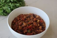 a white bowl filled with chili and beans next to some green leafy vegetables on a table