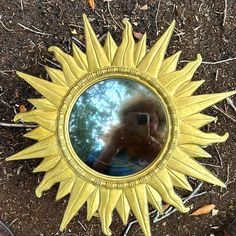 a sun shaped mirror sitting on the ground in front of some leaves and dirt with a person's reflection