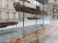 several wooden boats in a glass display case on the sidewalk outside an old building with cobblestone flooring