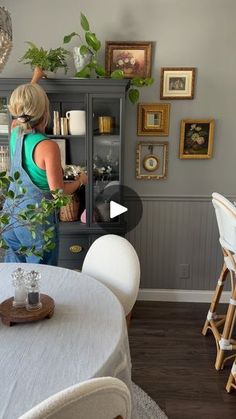 a woman standing in front of a book shelf filled with books and plants on top of a white table