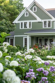 a green house with white trim and windows surrounded by flowers in the foreground, along with lots of greenery