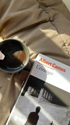 a person holding a coffee cup next to a book on a bed with a blanket