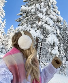 a woman with long blonde hair standing in front of a snow covered pine tree wearing ear muffs